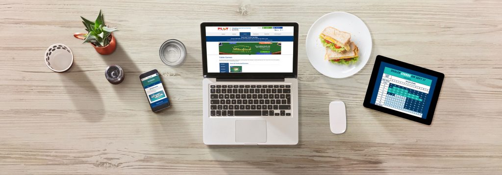 On top of a wooden table are a laptop with PlayOLG on the screen, a mobile phone with PlaySmart.ca on the screen, a tablet showing a Blackjack Strategy Card, a glass of water and a sandwich.