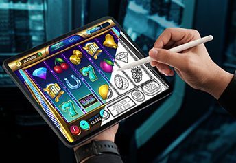 Against a casino backdrop, a hand is holding a tablet pen and drawing on a tablet which shows both fully rendered and rough sketches of slot machine game icons.