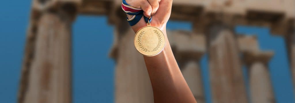 Hand holding gold medal in front of ancient Greek ruins.